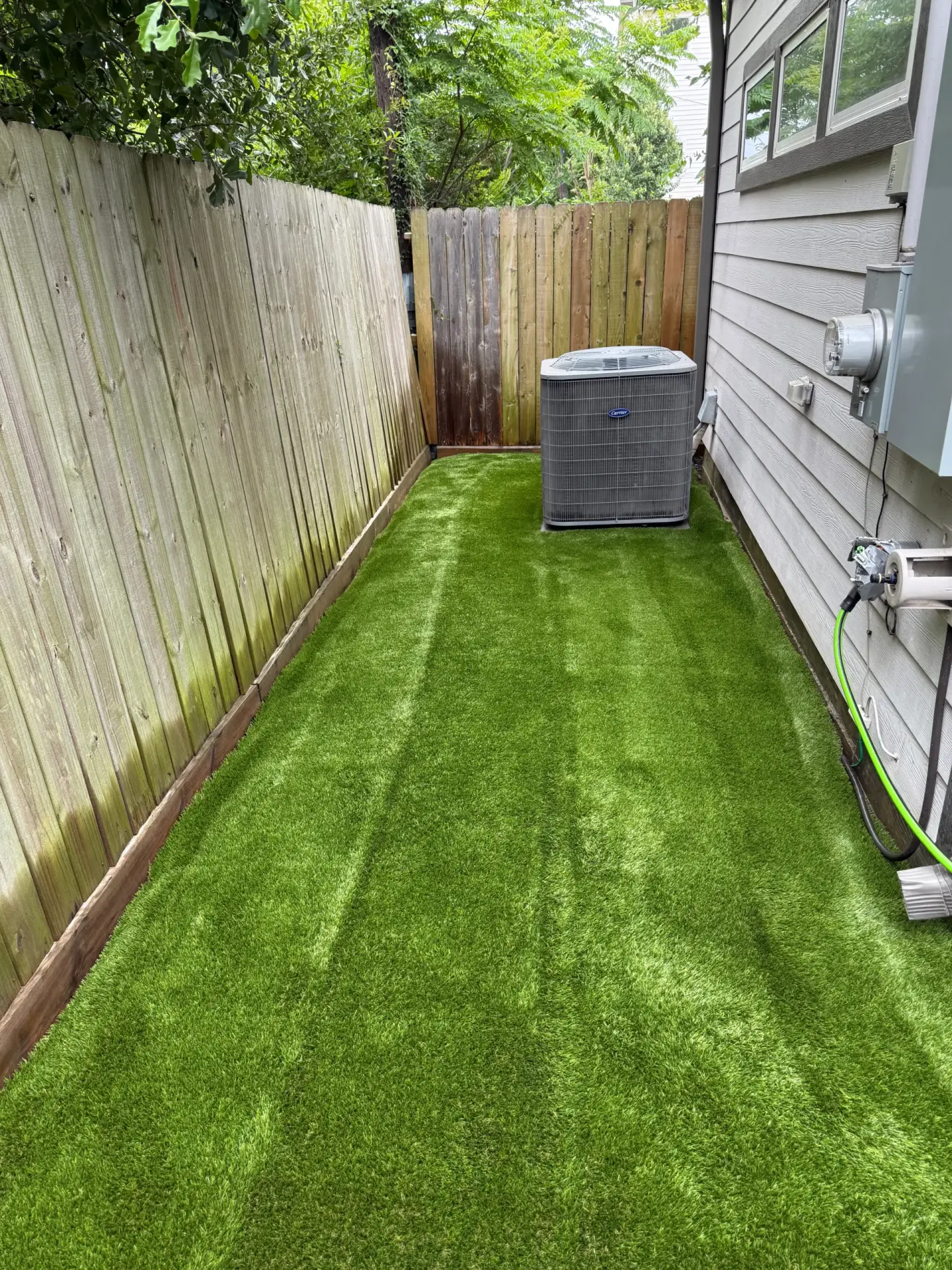 Fresh green artificial turf in a narrow side yard bordered by a wooden privacy fence and the exterior wall of a house, with an air conditioning unit and utility fixtures along the side.