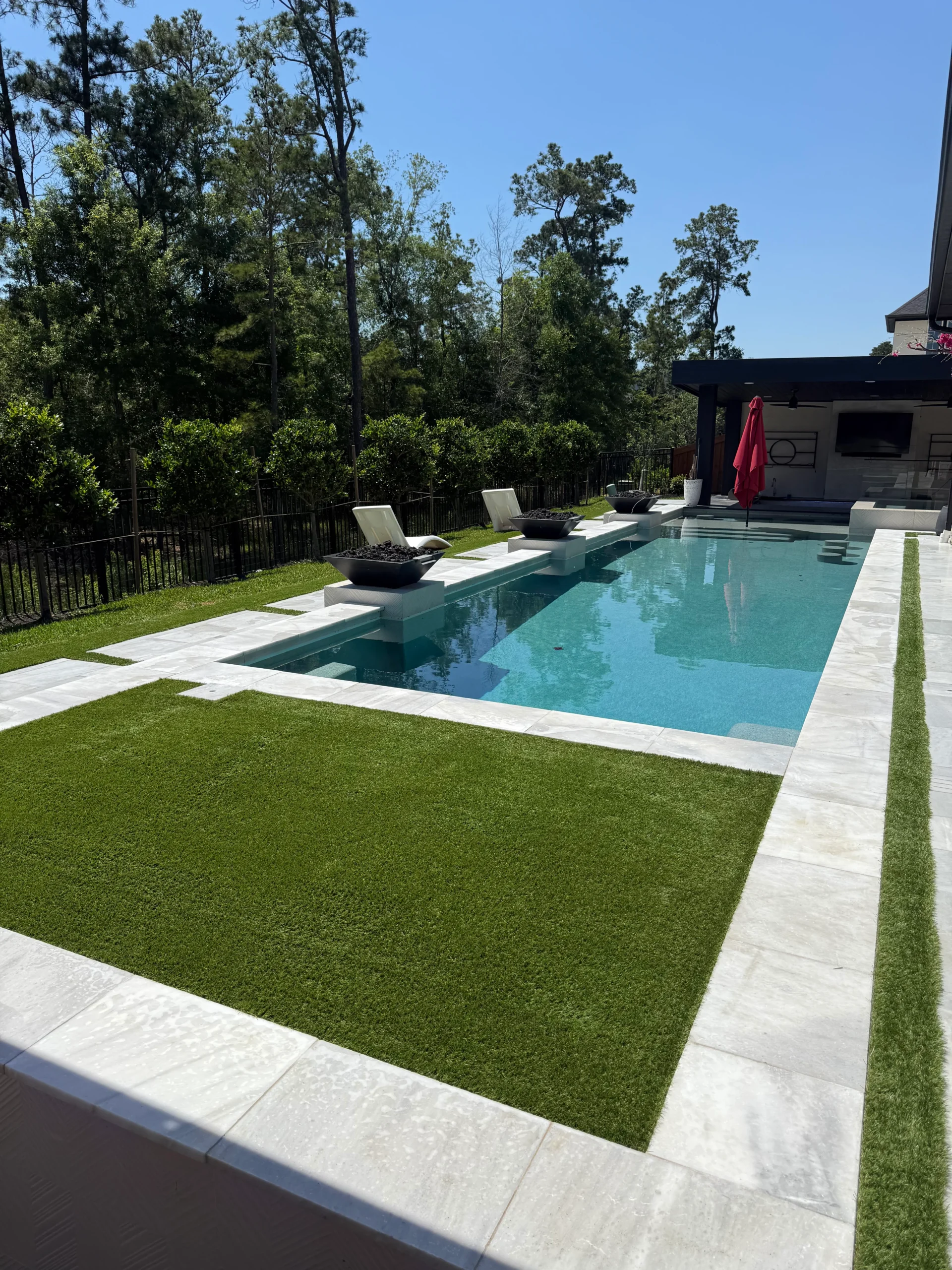 Luxury backyard with a rectangular swimming pool bordered by artificial turf and modern stone pavers, surrounded by landscaped greenery and mature trees under a clear blue sky.