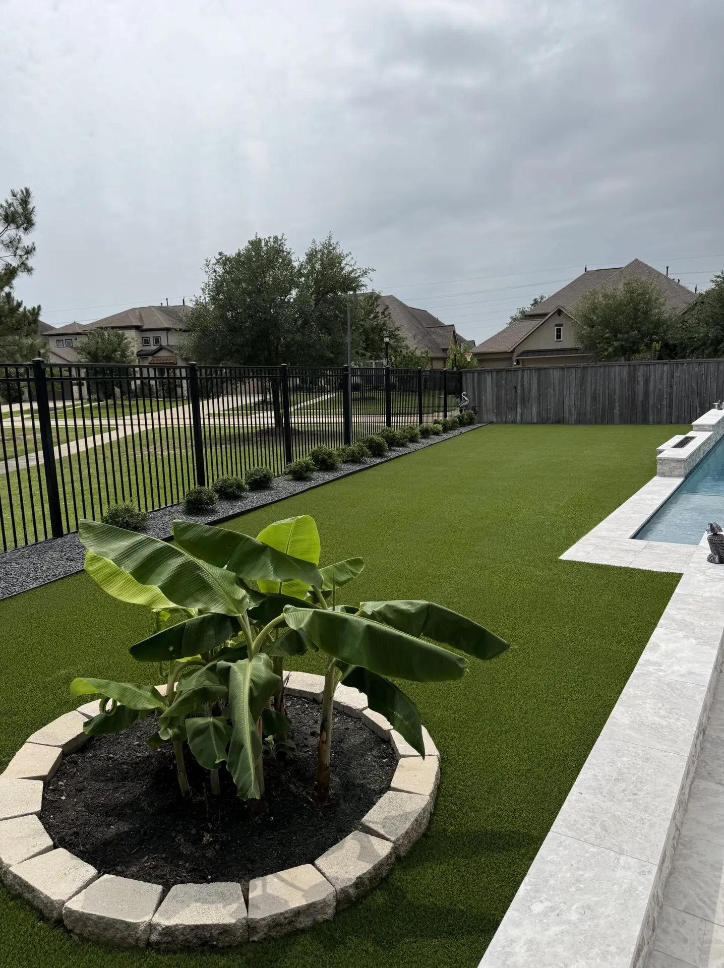 Residential backyard with a lush artificial turf lawn, a young tropical plant in a raised stone planter, and a swimming pool bordered by concrete decking under an overcast sky.