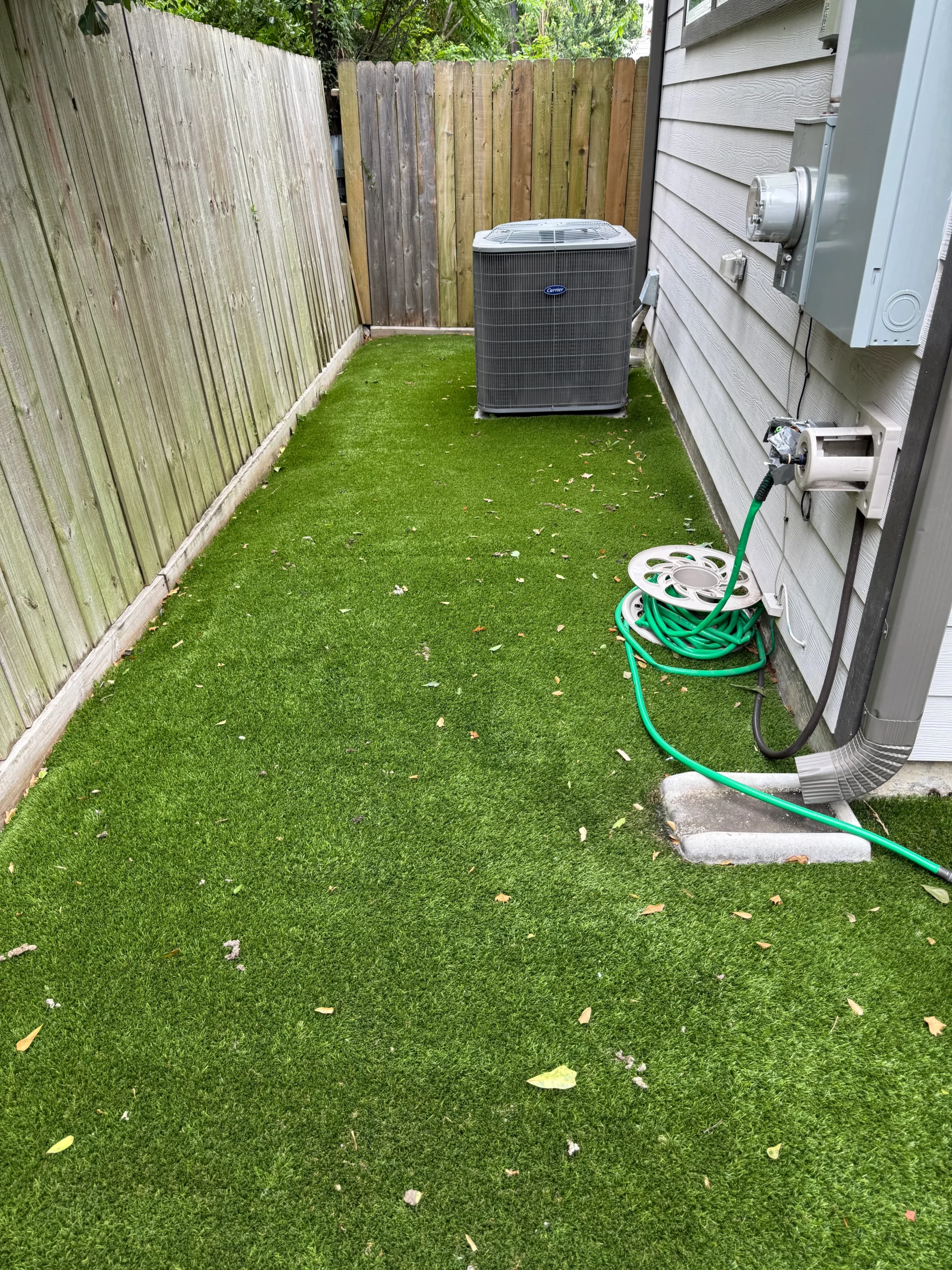 (Before) Houston Clean Dog Turf Artificial turf installed in a narrow side yard between a house and a wooden fence, with an outdoor air conditioning unit and utility equipment visible along the wall.