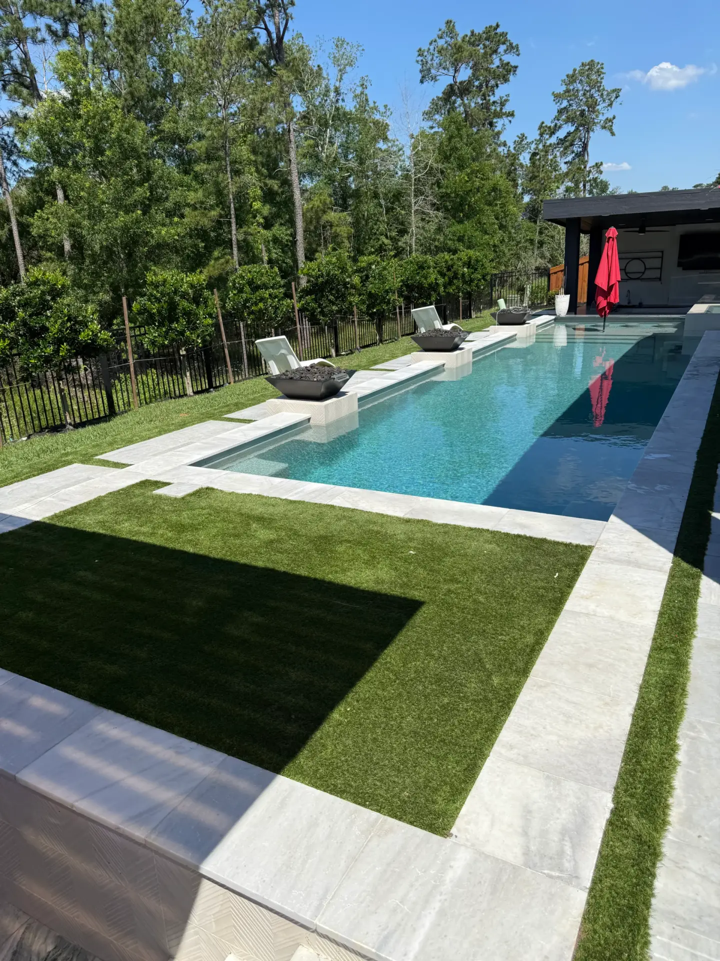 (Before) Houston Turf Clean Modern backyard featuring a rectangular swimming pool with a surrounding artificial turf lawn, light-colored paving, and a contemporary pool house set against a backdrop of trees.